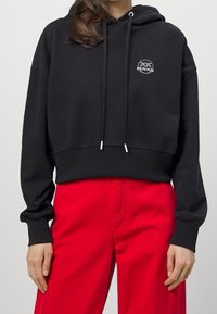 Svart kort hoodie med dragsko och en cirkulär logotyp på bröstet. Matchad med klara röda högmidjade byxor. Slät textur.