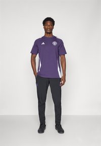 Paarse sportieve t-shirt met witte zijstrepen en logo, gecombineerd met zwarte broek met een embleem. Zwarte sneakers maken de outfit compleet.
