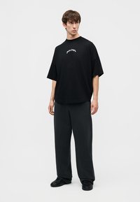 Noor mees seisab mustas ülepakutud Palm Angels T-särgis, lahtistes mustades pükstes ja mustades tennistes ühtlasel heledal taustal.