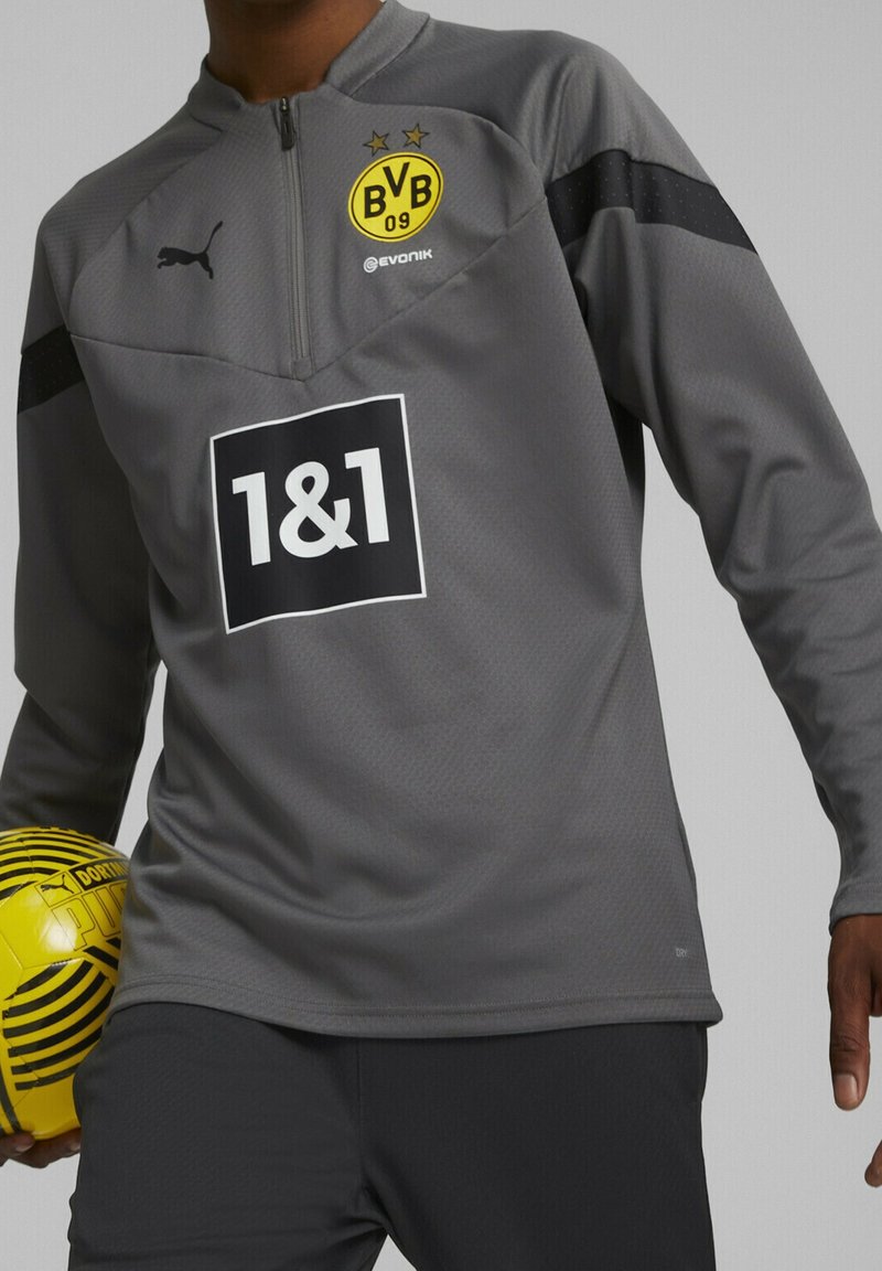 Harmaa vetoketjullinen urheilupaita mustilla yksityiskohdilla, jossa on logo ja musta suorakulmainen brändäys. Karkea kangas, yhdistetty keltaiseen ja mustaan raidalliseen palloon.