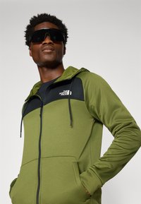 Olijfgroene hooded zipjacket met zwarte accenten, gemaakt van gladde stof, met verstelbare trekkoorden en voorzakken.