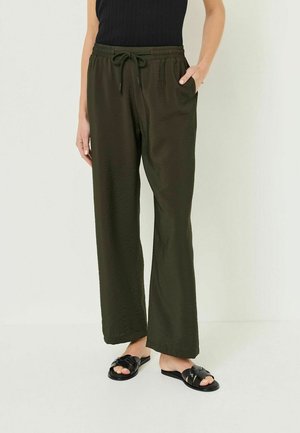 Kobieta ubrana w luźne ciemnozielone spodnie z troczkiem, czarny bezrękawnik i czarne plecione klapki, z jedną ręką w kieszeni.