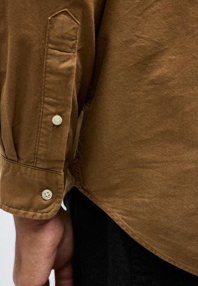 Καφέ μανσέτα πουκαμίσου με λευκά κουμπιά, φορεμένη στο αριστερό χέρι, σε αντίθεση με μαύρο παντελόνι στο φόντο.