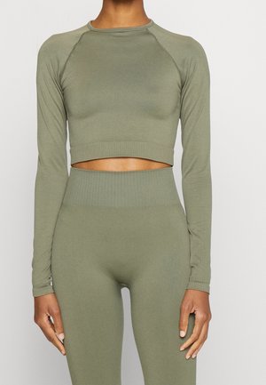 Vrouw draagt een olijfgroene cropped sporttop met lange mouwen en bijpassende hoog getailleerde leggings, staand tegen een effen lichte achtergrond.