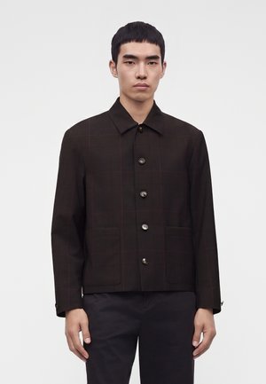 Jeune homme portant une veste marron foncé boutonnée avec deux poches avant et un pantalon noir sur un fond blanc uni.