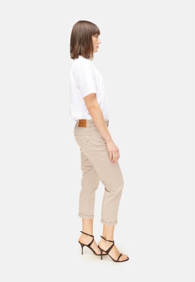 Donna in piedi di profilo, indossa una maglietta bianca, pantaloni beige corti e sandali neri con tacco alto e cinturini, su sfondo bianco.