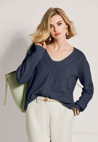 Blonde Frau, die einen marineblauen Kabelstrickpullover, cremefarbene Hosen mit einem dekorativen Gürtel, goldene Creolen, Armbänder und eine hellgrüne Umhängetasche trägt.