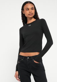 Čierny crop top s dlhým rukávom z mäkkého materiálu, s okrúhlym výstrihom a malým logom na prsiach, v kombinácii s čiernymi džínsami.