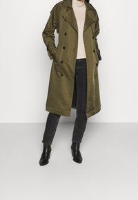 Imperméable trench coat vert olive avec boutons croisés, poches latérales et coupe décontractée, associé à un pull léger et un jean foncé.