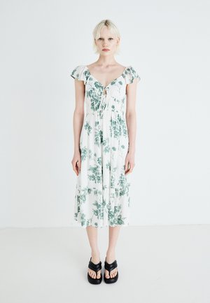 Vestido midi blanco con estampado floral verde, mangas cortas con volantes, cuerpo con cordón ajustable y dobladillo en capas. Llevado con sandalias de plataforma negras.