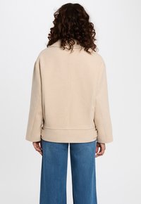 Persona con cabello rizado, vistiendo una chaqueta beige y jeans de pierna ancha azules, de pie con la espalda hacia el espectador frente a un fondo liso.