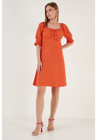 Orange dress with short puff sleeves, square neckline, and front ruching. Textured fabric, knee-length, paired with multi-colored sandals.