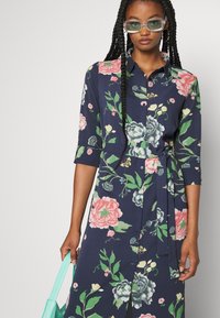Vestido camiseiro floral em azul marinho com padrões de flores em rosa e verde. Apresenta um colarinho, mangas três quartos, cintura com laço e fecho frontal com botões.
