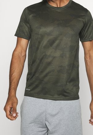 Man draagt een donker groen atletisch t-shirt met een hexagon-patroon camouflage en lichtgrijze korte broek, staat met ontspannen armen.