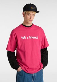 Lyserød bomulds-t-shirt med hvid tekst "fortæl en ven." lagdelt over en sort langærmet trøje. Blå denimbukser. Sort kasket med et logo.