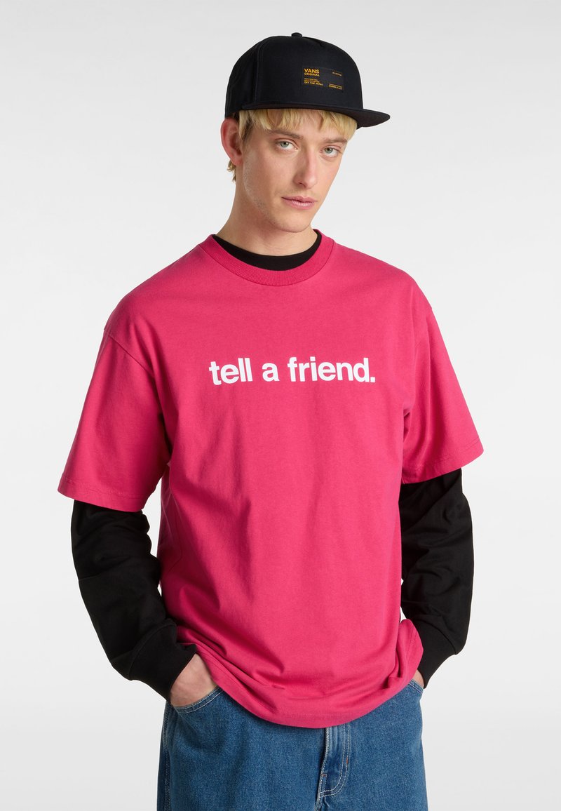 Lyserød bomulds-t-shirt med hvid tekst "fortæl en ven." lagdelt over en sort langærmet trøje. Blå denimbukser. Sort kasket med et logo.
