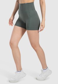 Vrouw in donkergroene high-waisted sportshorts met zijzakken en witte sneakers, staand met de benen iets uit elkaar tegen een lichte achtergrond.