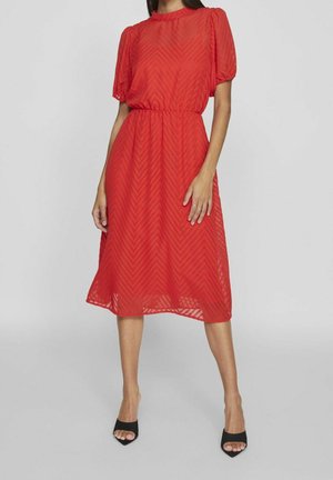 Femme portant une robe rouge longueur genoux avec des manches courtes bouffantes et un motif chevron, associée à des talons ouverts noirs.