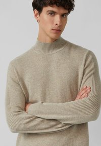 Mann mit verschränkten Armen, der einen beigen, gerippten Stehkragenpullover trägt und leicht zur Seite schaut, vor einem einfarbigen, hellgrauen Hintergrund.