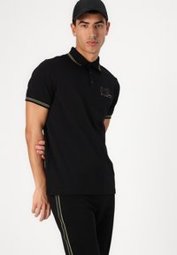 Schwarzes Polohemd mit gelben Akzenten und Logo-Patch, kurze Ärmel, mit Kragen, kombiniert mit passenden schwarzen Hosen mit seitlichem Streifen.