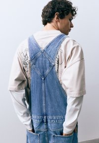 Jeans tuinbroek in lichtblauw met verstelbare bandjes, opgestikte zakken en een losse pasvorm, gedragen over een beige t-shirt met print en een witte lange mouw.