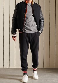 Chaqueta varsity negra con mangas de cuero, camiseta gris, pantalones de chándal negros y zapatillas blancas con calcetines a rayas rojas y negras.