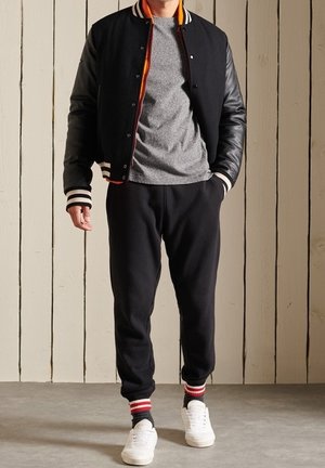 Veste universitaire noire avec des manches en cuir, t-shirt gris, pantalon de survêtement noir et baskets blanches avec des chaussettes rayées rouges et noires.