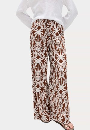Brune bukser med vide ben med hvide indviklede blomster- og palmemønstre, båret med en hvid top og sandaler med åbne tæer.