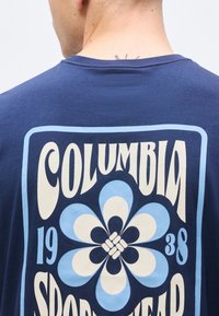 Πίσω μέρος ατόμου που φορά μπλούζα Columbia Sportswear σε ναυτικό μπλε χρώμα με μεγάλο ρετρό λουλούδι και κείμενο "1938" σε μπλε και εκρού αποχρώσεις.