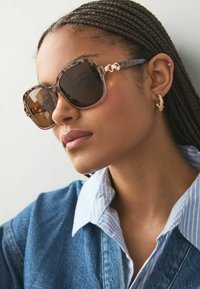 Übergroße Sonnenbrille mit einem braunen Schildpatt-Rahmen, dunklen Gläsern und goldfarbenen Akzenten. Tragen Sie sie zu einer Jeansjacke und einem gestreiften Kragenhemd.