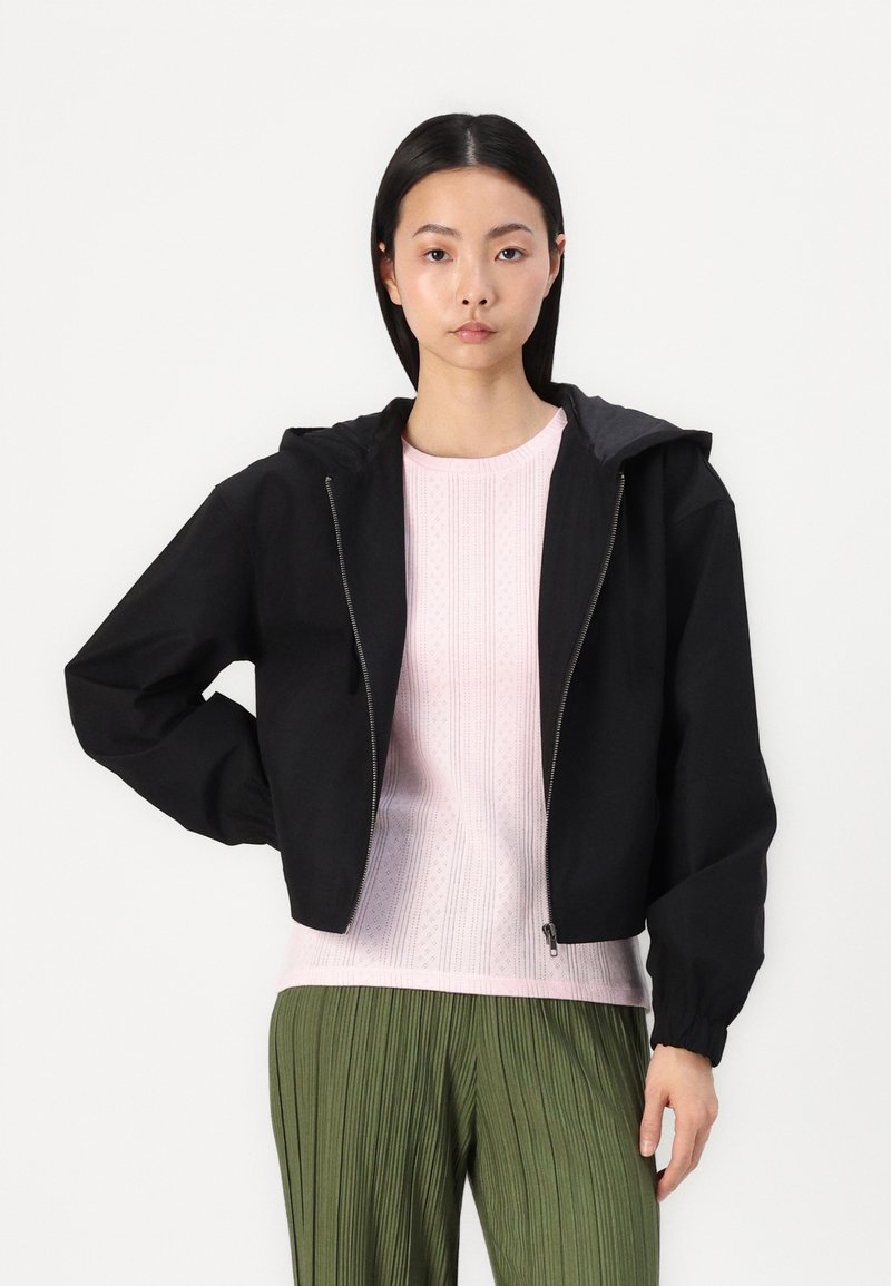 Giacca nera corta con cappuccio, chiusura con zip e polsini elasticizzati, indossata sopra un maglione rosa chiaro a trama e pantaloni green plissettati.