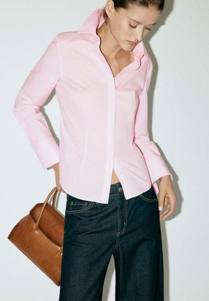 Femme portant une chemise rose clair boutonnée et un jean foncé, tenant un sac à main en cuir marron dans sa main gauche sur fond blanc.