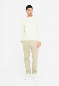 Svetlo kremna majica z reliefnim logotipom, kombinirana z bež hlačami. Obleko zaključujejo bele superge, ki poudarjajo sproščen kroj in gladko teksturo.