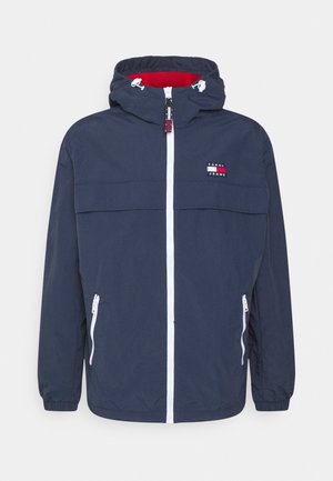 Man draagt een marineblauwe capuchonjas met witte ritssluitingen en een Tommy Jeans-logo op de borst, gecombineerd met een beige broek.