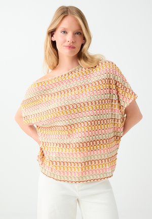 Donna bionda con capelli sciolti indossa un top colorato all'uncinetto con motivi beige, rosa, gialli e marroni e pantaloni bianchi su sfondo bianco.