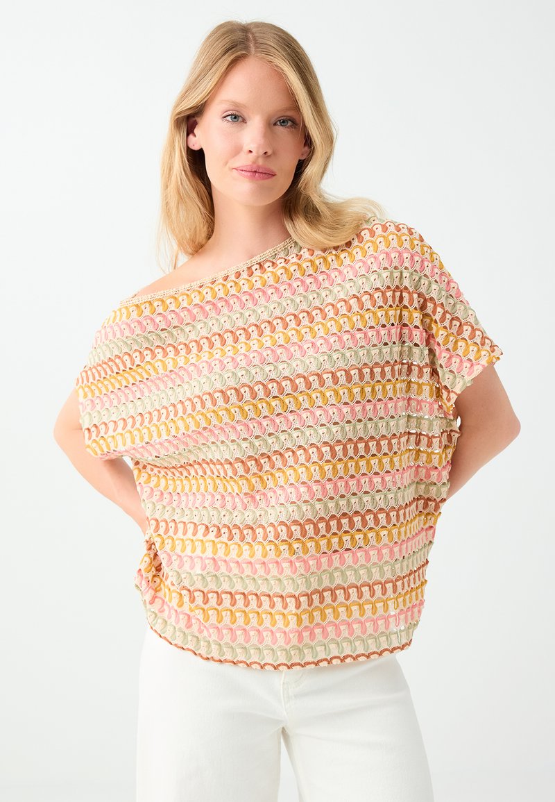 Femme blonde aux cheveux lâchés porte un haut en crochet coloré avec des motifs beige, rose, jaune et marron ainsi qu'un pantalon blanc sur fond blanc.