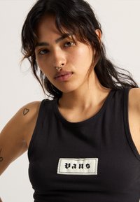 Canotta nera con una patch rettangolare bianca con la scritta "vans" in lettere nere. Il tessuto ha una texture liscia e un design aderente.