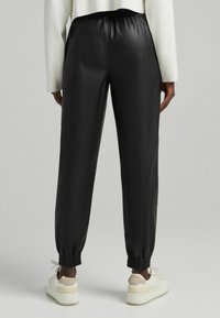 Schwarze Sweatpants aus Kunstleder mit elastischem Bund und Bündchen. Glatte Textur, gerades Bein. Kombiniert mit weißen Sneakers.