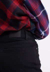 Svarta denimjeans med hög midja och lätt stretch, med bakfickor och en etikett vid midjebandet. Kombineras med en röd och blå rutig skjorta.