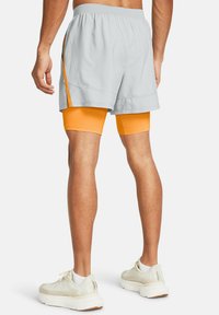 Grå atletiske shorts over orange kompressionshorts, med en mesh sidelomme. Modellen har hvide sneakers med teksturerede detaljer på.