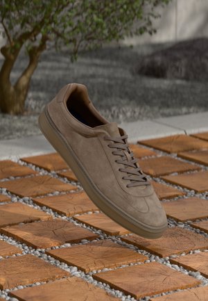 Lysebrun ruskindssneaker med snørebånd, der svæver over en træflisebelagt gulv udendørs nær et lille træ.