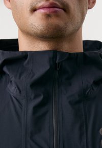 Veste à capuche noire avec un col haut, dotée d'une fermeture éclair et d'un tissu texturé. Les détails incluent des lignes de couture et un design minimaliste.
