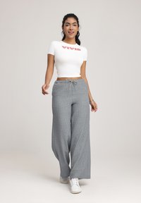 Witte top met korte mouwen met "VIVID" in het rood, gecombineerd met grijze brede broek met een gestreept design en een trekkoord. Witte sneakers.