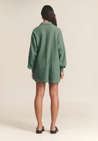 Zelené, textúrované šaty košeľového strihu s dlhými rukávmi, bočnými vreckami a gombíkovými manžetami. Tkanina vyzerá ľahká a uvoľnená.