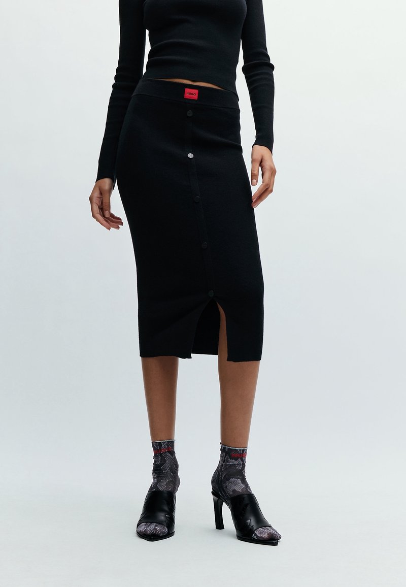 Femme portant une jupe midi noire à boutons avec une fente sur le devant, un haut ajusté noir à manches longues, des chaussettes à motif serpent, et des chaussures à talons noires.