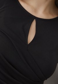 Abito nero realizzato in tessuto morbido, con scollatura a chiave e dettagli arricciati sul lato, che creano una silhouette aderente.