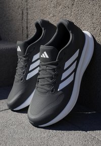 Black athletic shoes made of mesh material with three white stripes on the sides and textured soles, featuring laces and rounded toes.