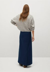 Donna con lunghi capelli castani che indossa un maglione grigio lavorato a maglia e una lunga gonna blu navy, in piedi con le spalle rivolte verso la macchina fotografica.