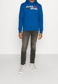 Modrá mikina s kapucí s logem "JACK&JONES", přední kapsa, v kombinaci s tmavě šedými džínami a bílými vysokými teniskami.