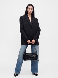 Femme portant un blazer noir oversize et un jean à jambes larges tenant un sac à main noir avec des cheveux noirs raides sur un fond uni.